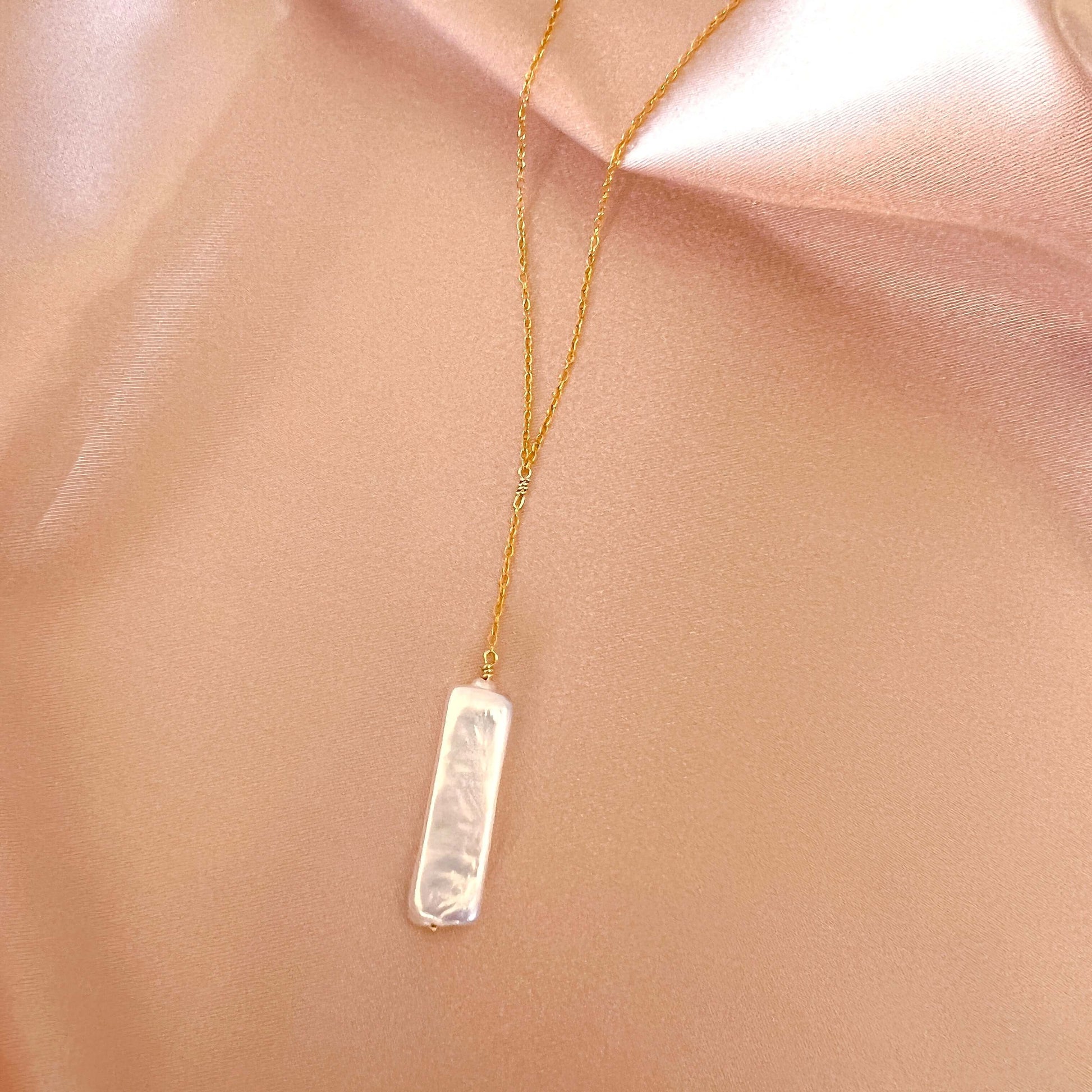 Gold necklace with a rectangular pendant on a soft pink fabric background