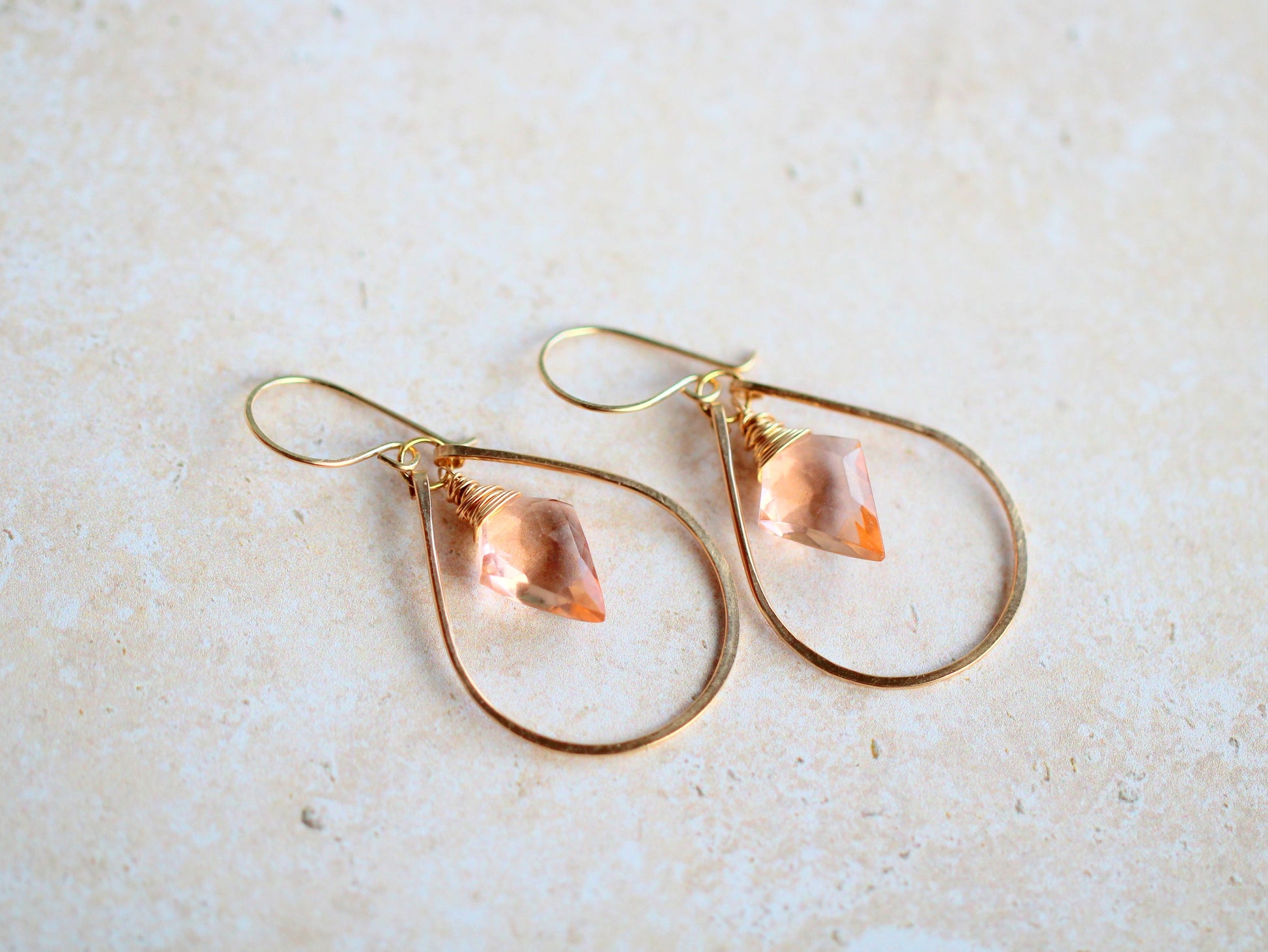 Gold hoop earrings with teardrop-shaped pink gemstones on a light beige background