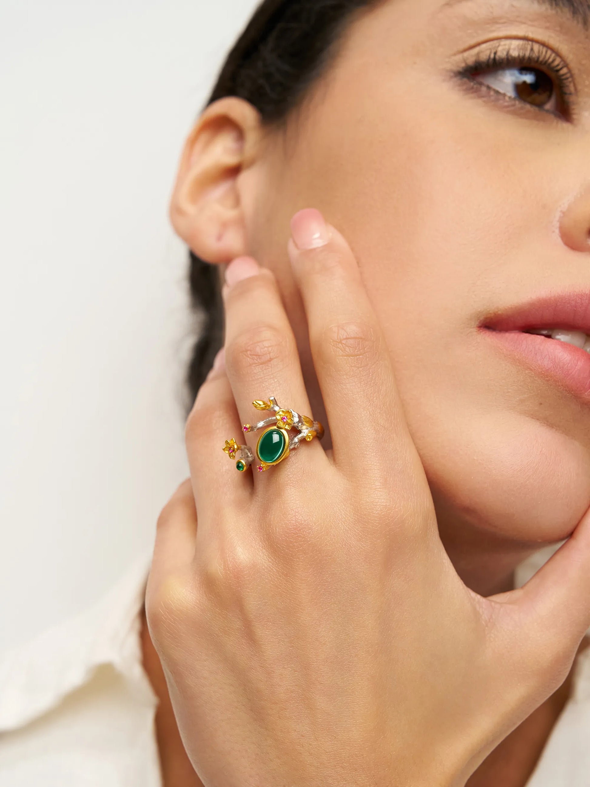 Close-up of a hand wearing a gold ring with a green gemstone, touching the face.